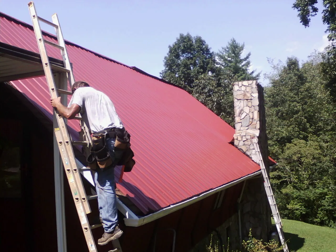 Roofer on a ladder beside a red metal roof and stone chimney — Soffit & Fascia Repair by Roofing Brothers in Pond Creek